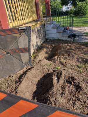 Two stumps near the foundation and porch have already been ground down. Two holes remain where the stumps used to be, next to the foundation and porch.